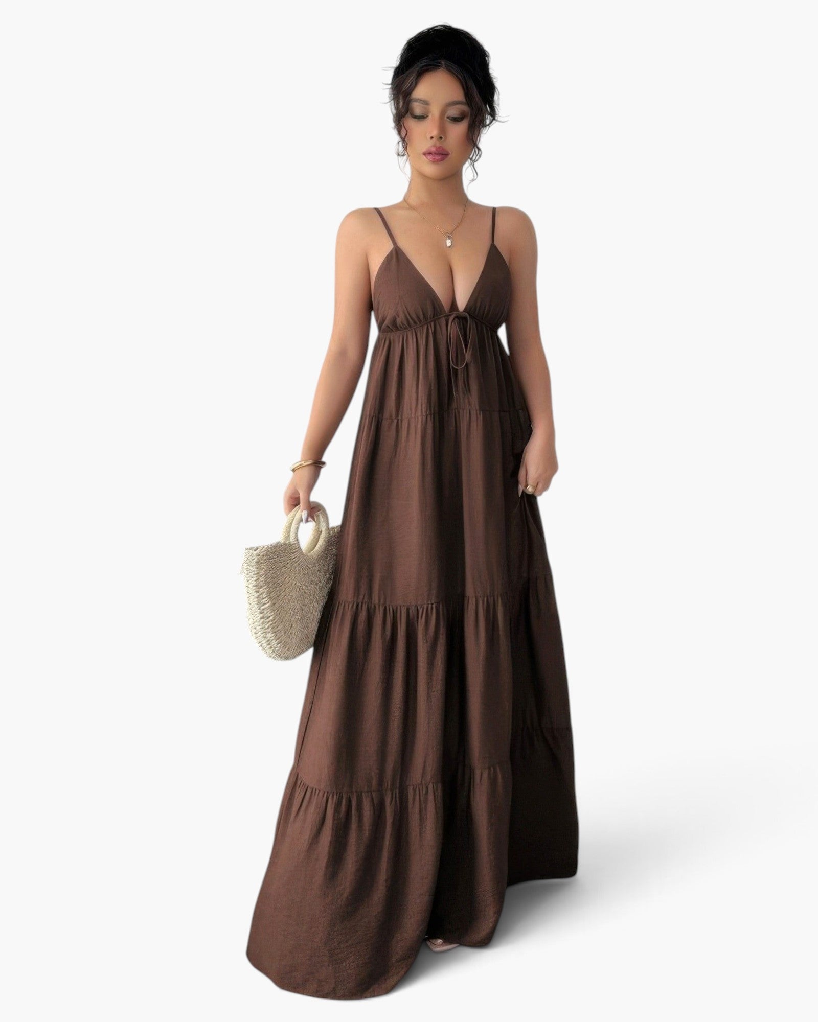 A woman modeling a brown tiered maxi dress with thin spaghetti straps and a drawstring tie at the bust, holding a small woven straw handbag against a plain light gray studio background.