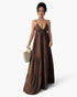 A woman modeling a brown tiered maxi dress with thin spaghetti straps and a drawstring tie at the bust, holding a small woven straw handbag against a plain light gray studio background.
