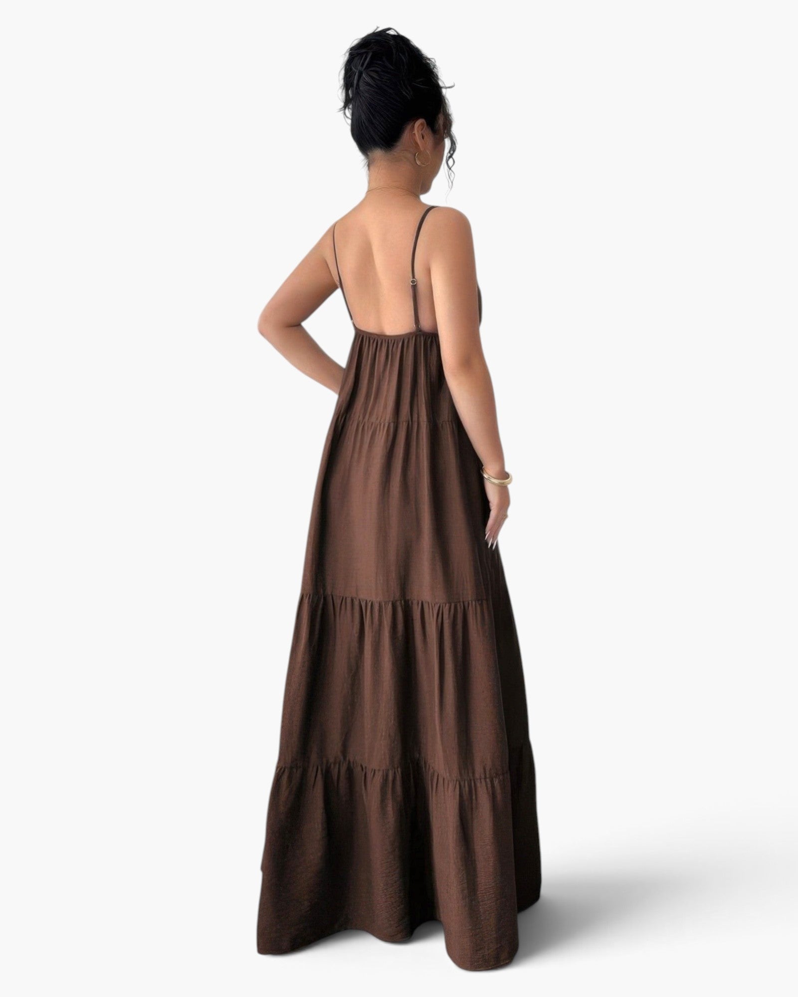A woman viewed from the back wearing a brown tiered maxi dress with thin spaghetti straps and an open back, photographed against a clean white studio background.
