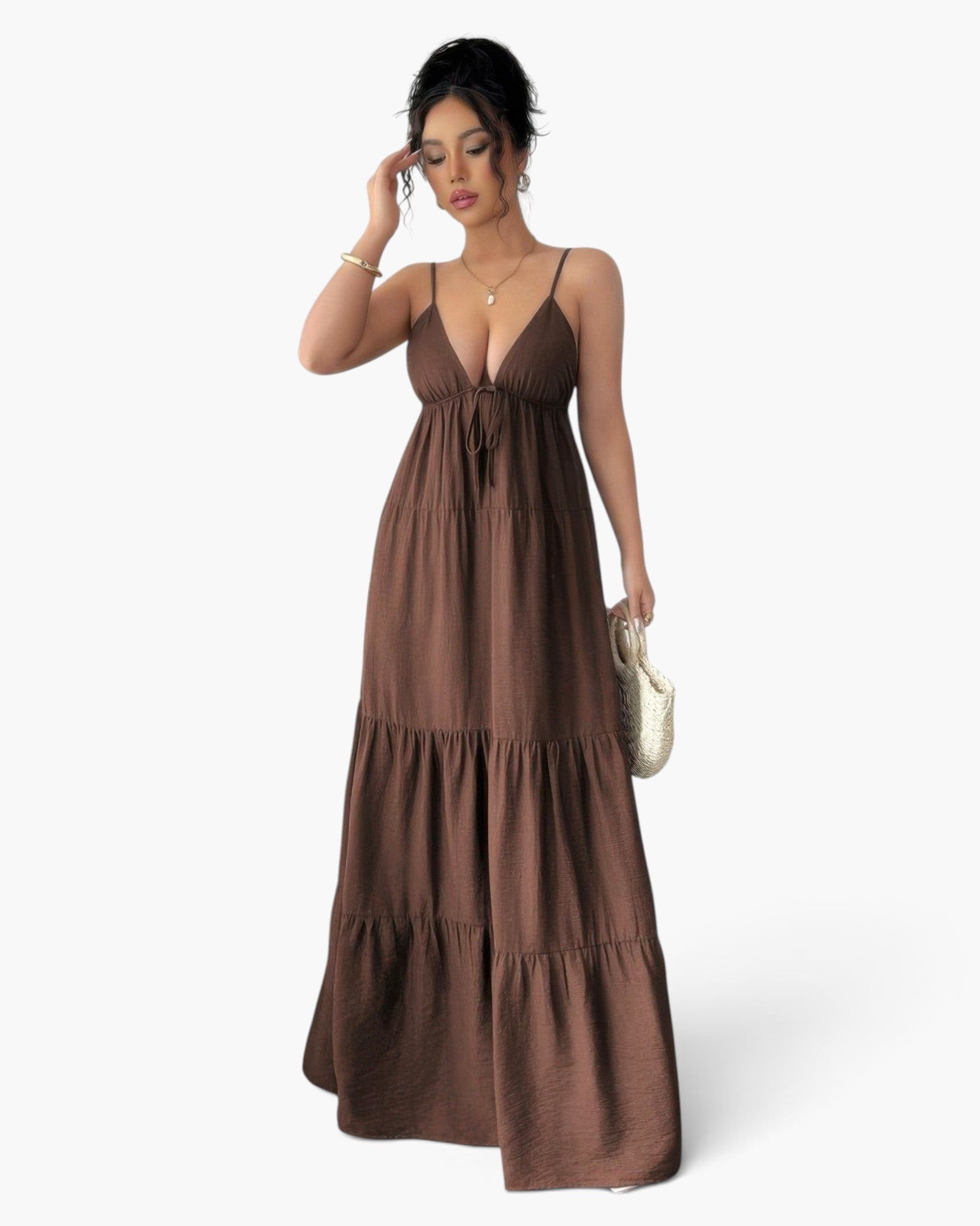 A woman modeling a brown tiered maxi dress with spaghetti straps and a tie-front waist, standing against a light gray studio background and holding a small woven clutch.