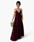A woman modeling a long, deep burgundy maxi dress with thin spaghetti straps, a V-neckline, and a tiered skirt, holding a small beige woven handbag against a plain white background.