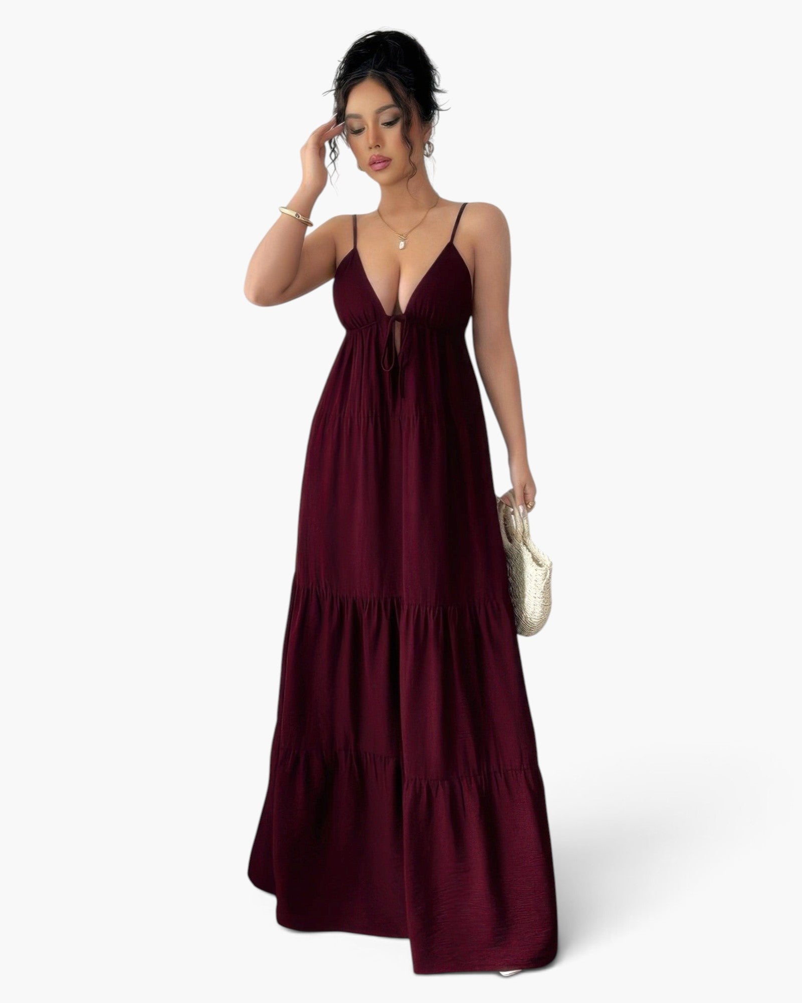 A woman modeling a long, tiered maxi dress in deep burgundy with thin spaghetti straps and a V-neckline, holding a small white woven clutch against a light gray studio background.