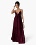 A woman modeling a long, tiered maxi dress in deep burgundy with thin spaghetti straps and a V-neckline, holding a small white woven clutch against a light gray studio background.