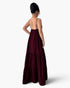 A woman shown from the back wearing a long, dark burgundy maxi dress with thin spaghetti straps and a tiered skirt, set against a plain light gray studio background.