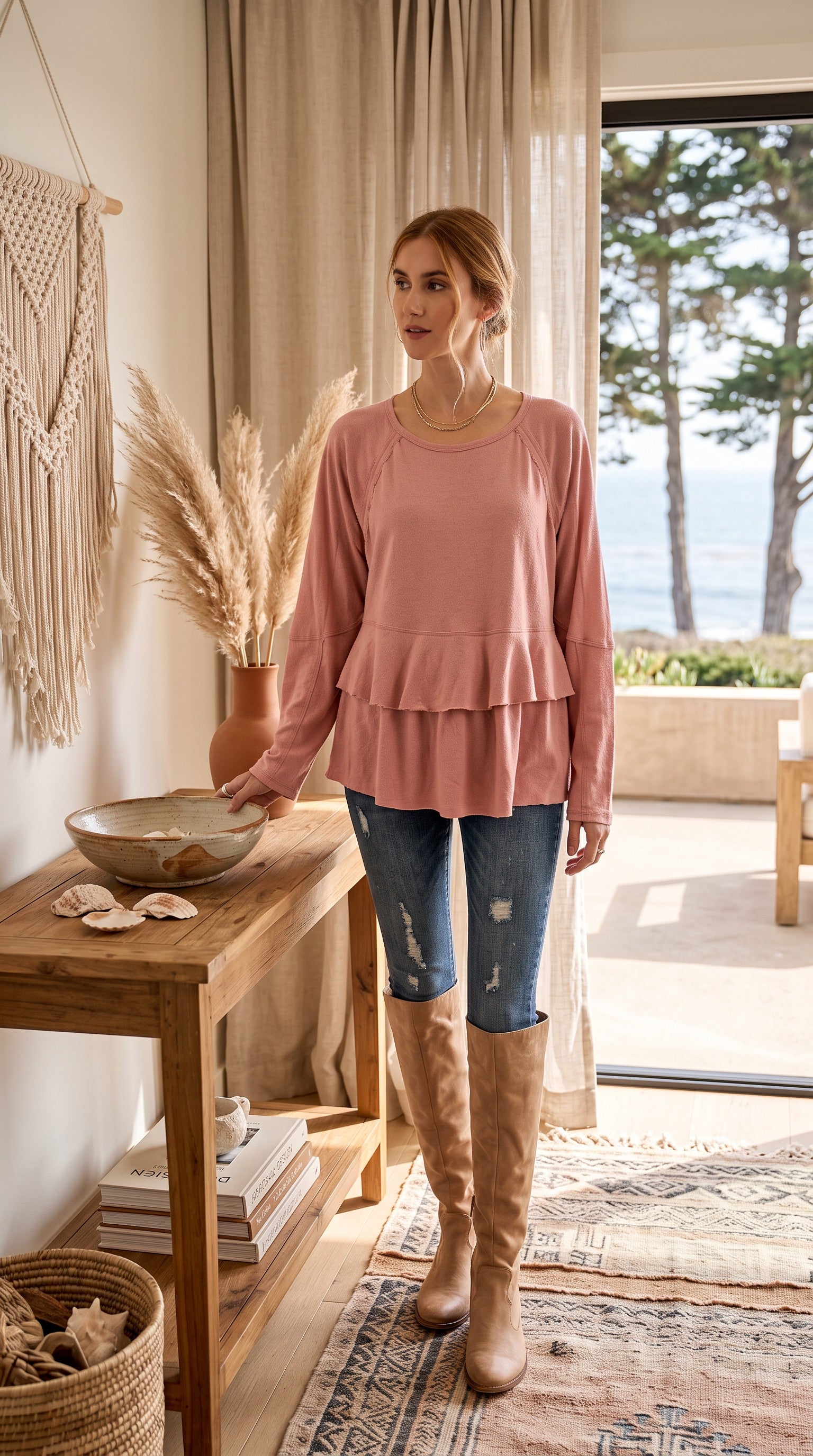 A woman stands in a bright, modern room wearing a dusty rose long-sleeve ruffled top, blue distressed jeans, and tan knee-high boots. A wooden console table, pampas grass, and a window overlooking trees and water are visible in the background.