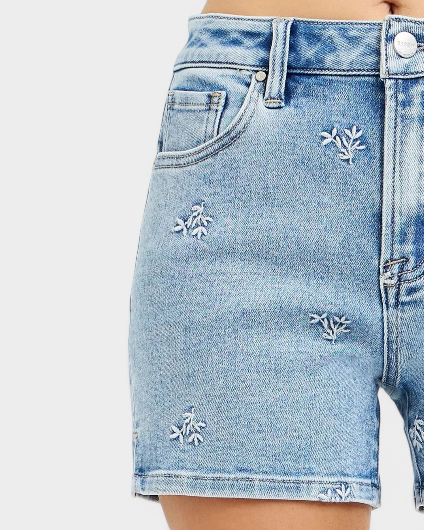 Beach Rose Co. light wash denim shorts with white embroidered floral details, high-rise fit, and short length.