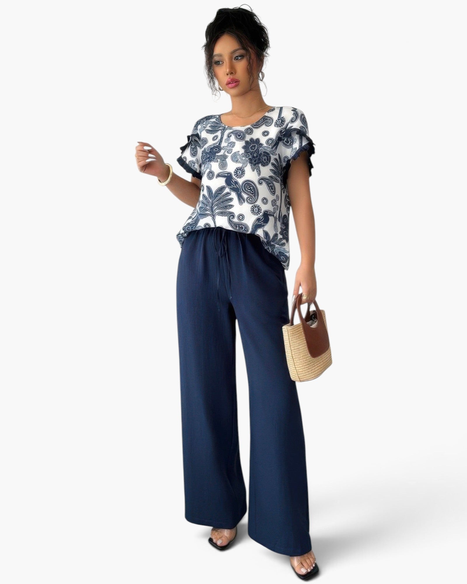 A woman modeling a short-sleeve ruffled blouse with a navy and white paisley pattern, paired with navy wide-leg drawstring pants and holding a small woven handbag, against a light gray studio background.