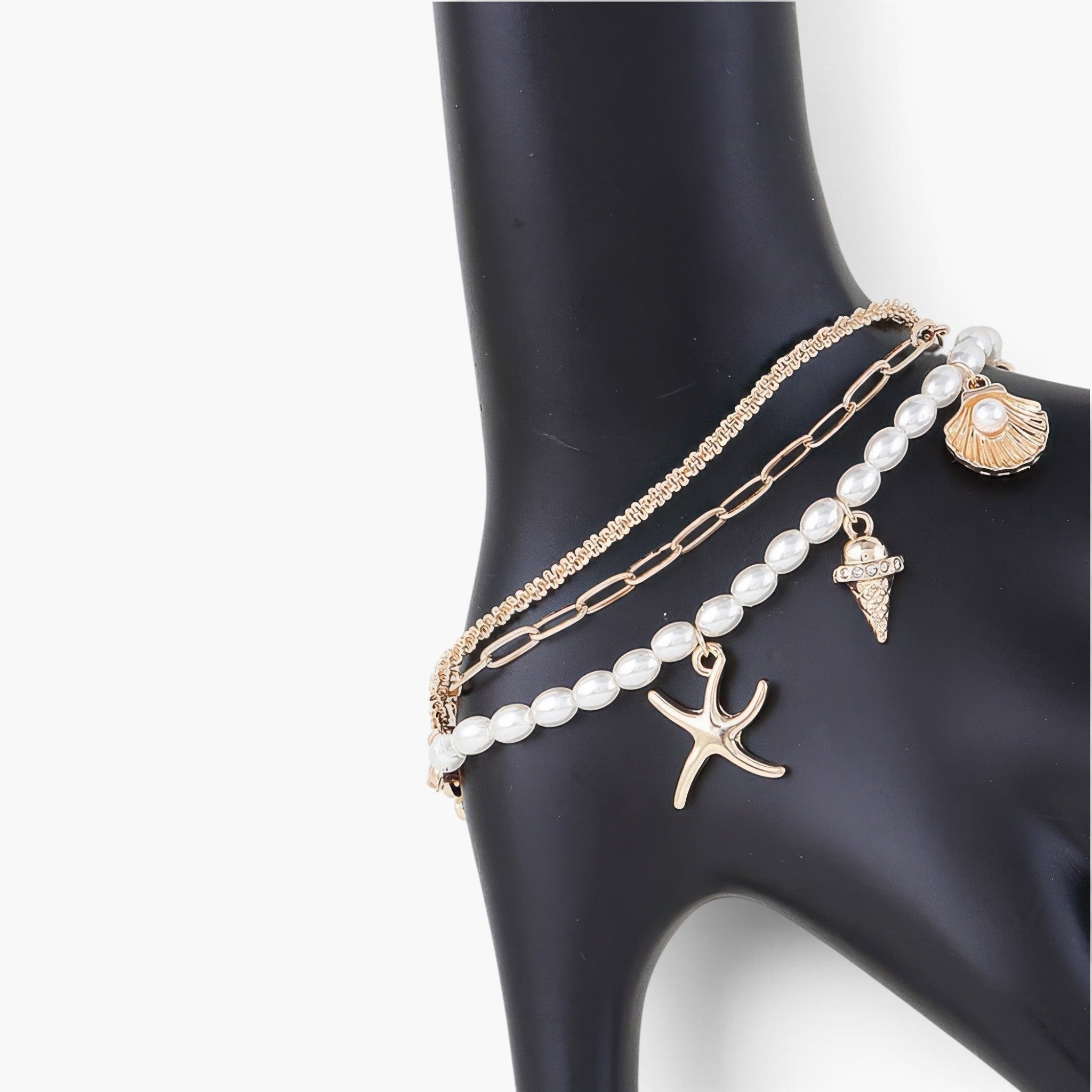 Beach Rose Co. layered gold chain bracelet featuring pearl-like beads, starfish, and seashell charms on a black stand.