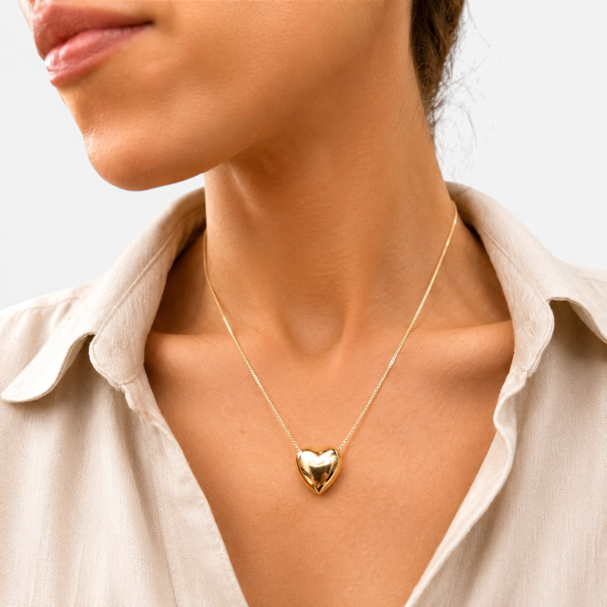 Beach Rose Co. gold puff heart pendant necklace worn on a delicate chain by a woman in a beige linen shirt.
