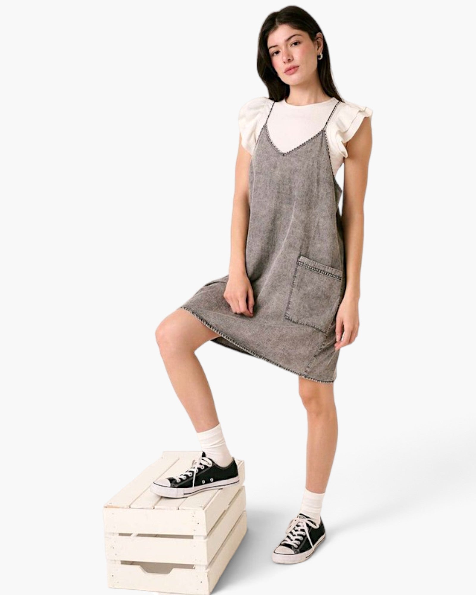 Beach Rose Co. gray washed denim overall dress with oversized pockets and slender straps worn over a white flutter top.