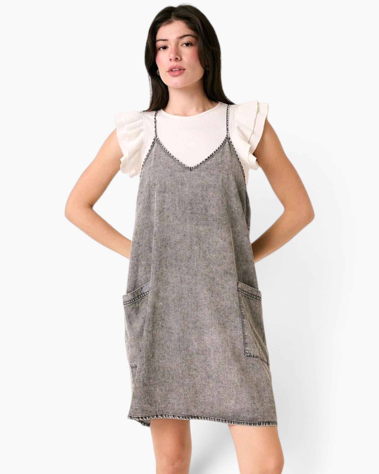 Model wearing a Beach Rose Co. gray washed denim overall dress with oversized pockets over a white flutter-sleeve top.