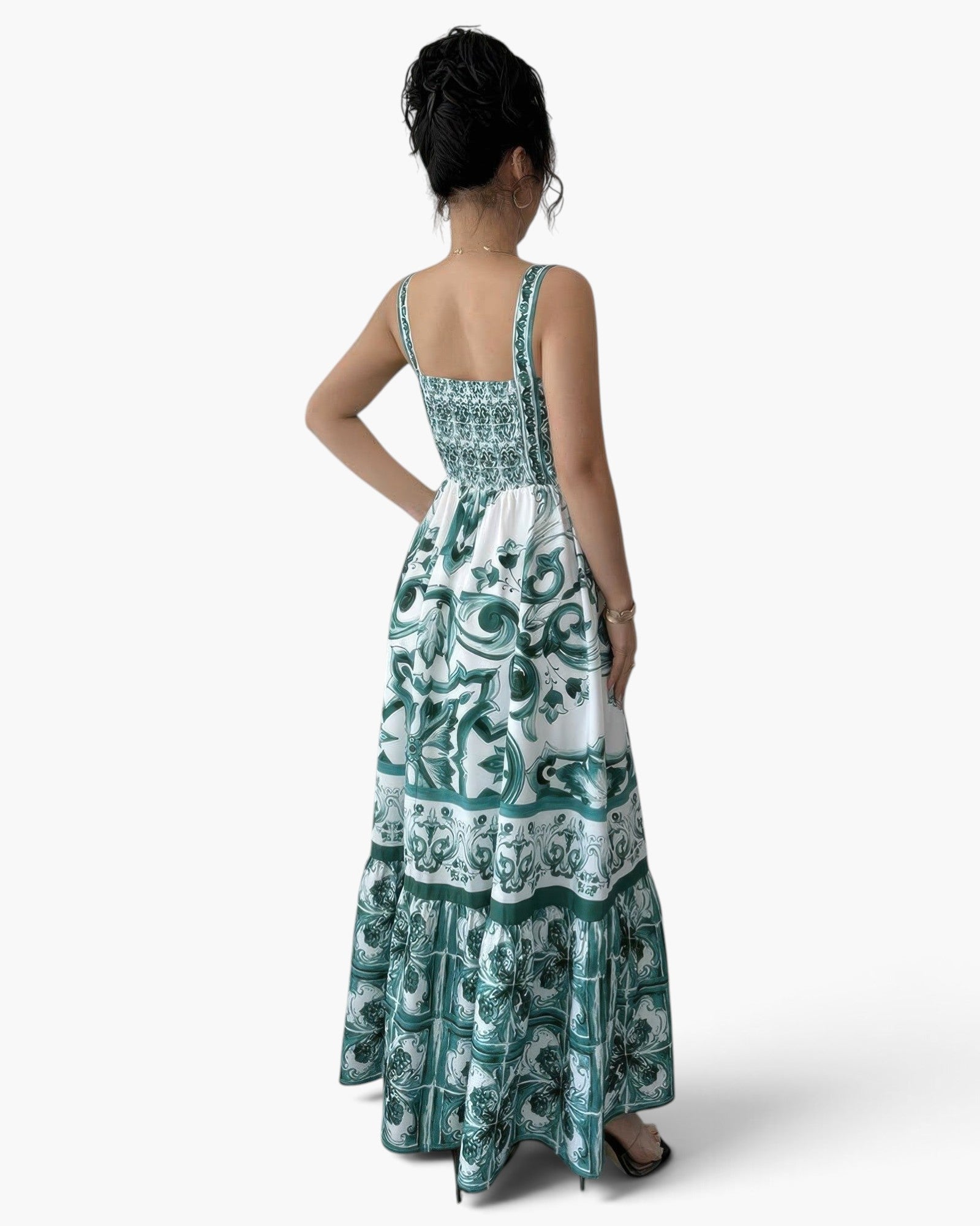 A woman shown from the back wearing a sleeveless maxi dress with a white base and teal green ornate pattern, photographed against a clean white studio background.