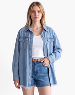 Beach Rose Co. light wash denim shirt with a frayed hem, pointed collar, and chest pockets worn over a white tank top.