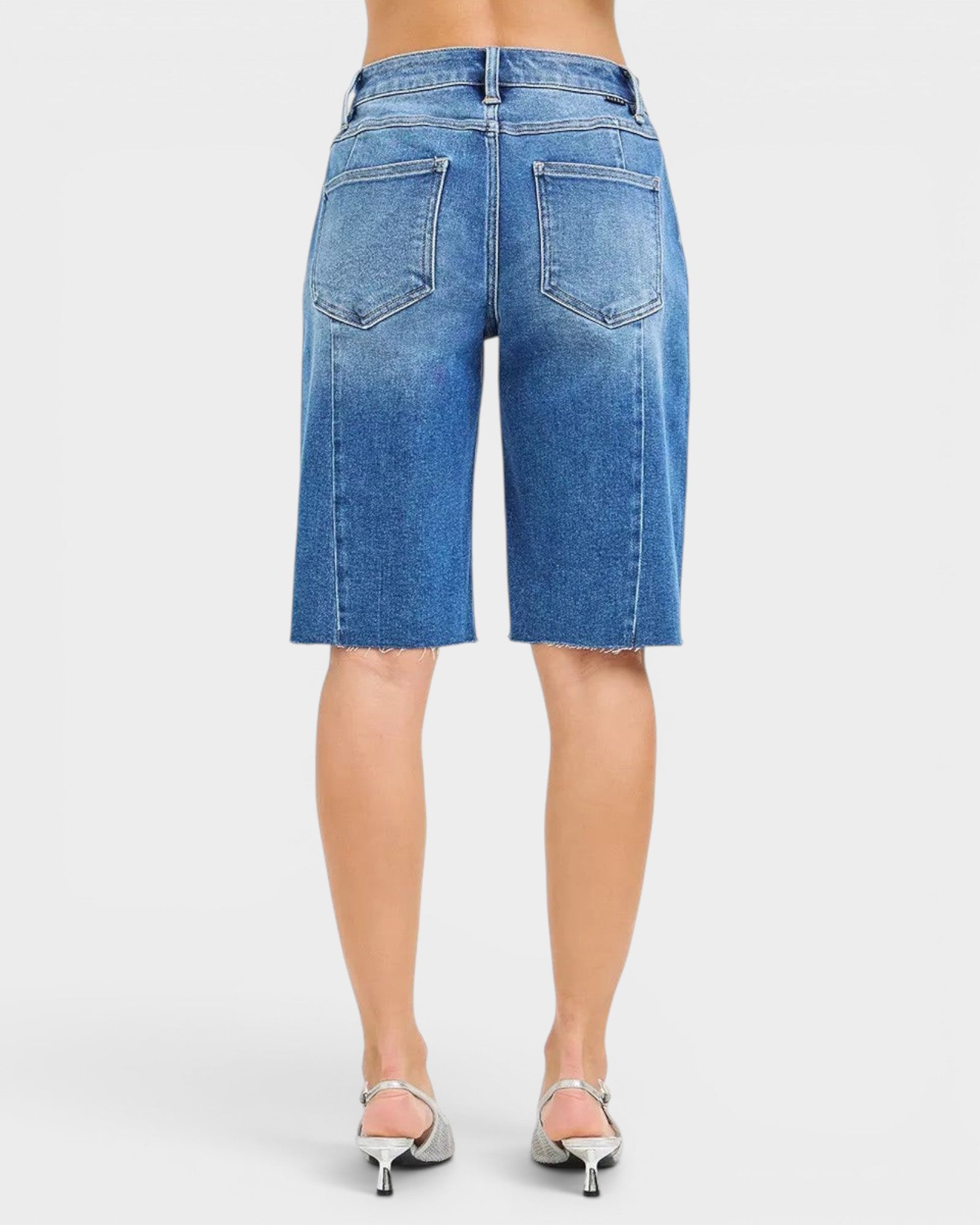 Beach Rose Co. Medium wash denim Bermuda shorts with raw hem and five-pocket styling. Made from a cotton blend with stretch.