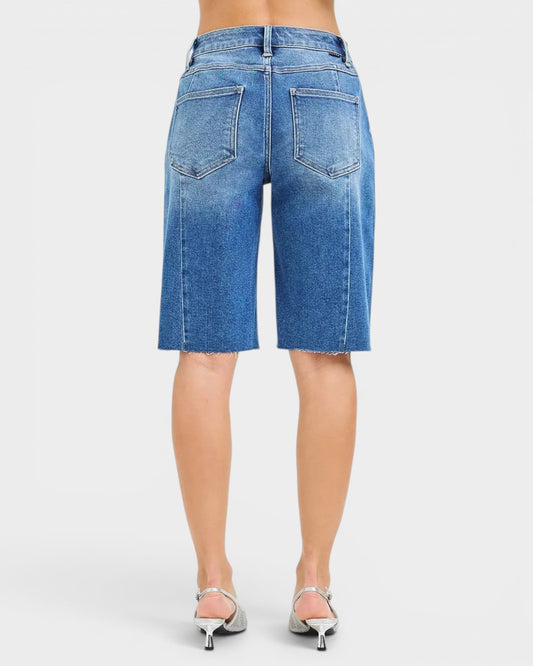 Beach Rose Co. Medium wash denim Bermuda shorts with raw hem and five-pocket styling. Made from a cotton blend with stretch.
