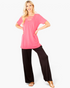 Beach Rose Co. neon pink V-neck tunic top with strappy neckline detail worn by a blonde model with black trousers.