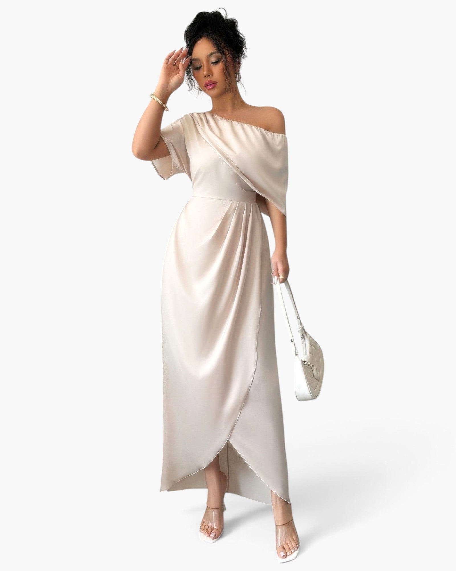 A woman modeling a floor-length off-the-shoulder draped dress in a light champagne beige satin fabric, paired with a white handbag and clear high-heeled sandals, set against a plain white studio background.