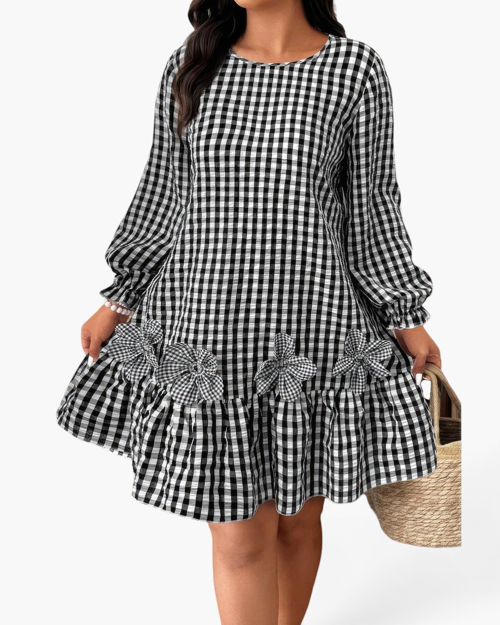 A woman wearing a black and white gingham long-sleeve dress with a ruffled hem and matching fabric flower appliqués, holding a woven straw bag against a plain light background.
