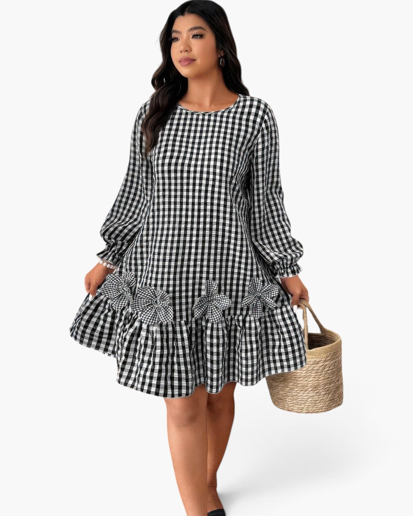 A woman modeling a black and white gingham long-sleeve dress with ruffled trim and a matching gingham bow, holding a woven straw basket bag against a light gray studio background.