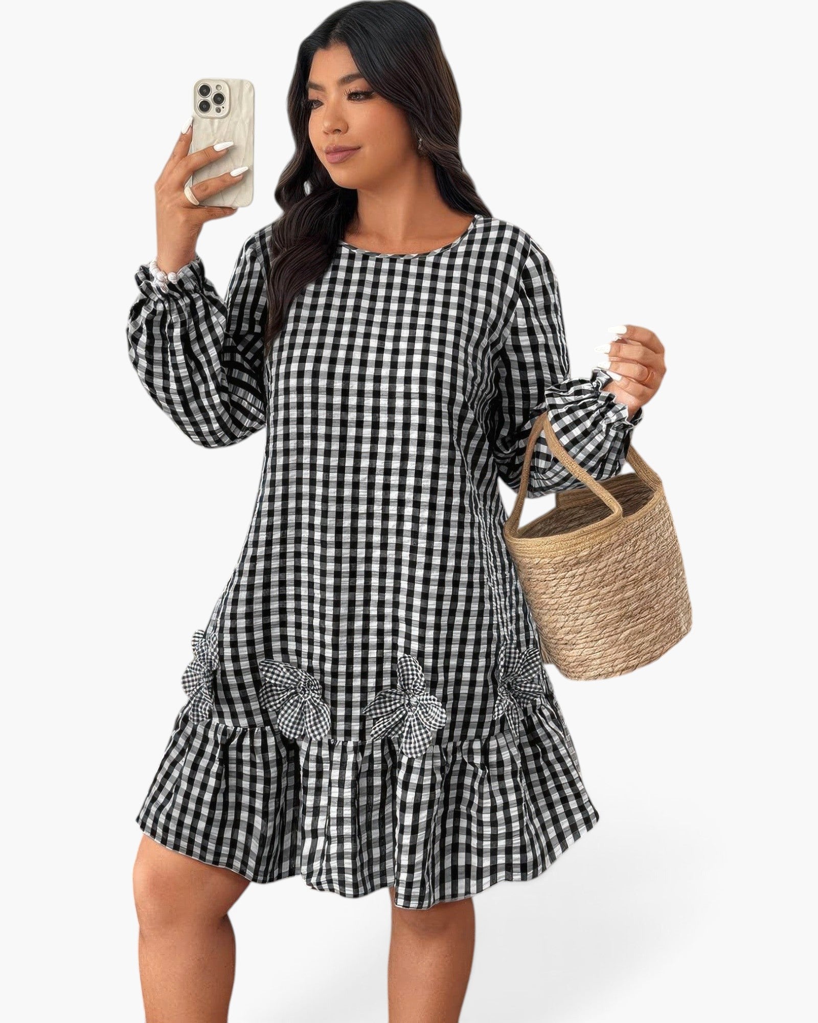 A woman takes a mirror selfie wearing a black and white gingham long-sleeve mini dress with a ruffled hem and decorative bow details. She is holding a woven straw tote bag against a clean white background.
