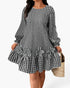 A woman wearing a black and white gingham long-sleeve dress with a ruffled hem and matching fabric flower appliqués, holding a woven straw bag against a plain light background.