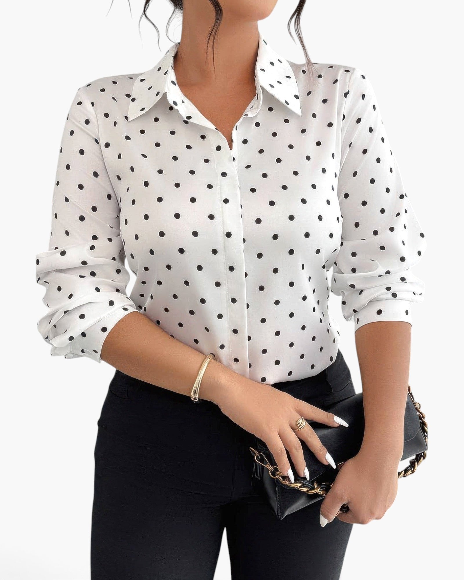 A woman wearing a white long-sleeve button-up blouse with black polka dots, paired with black pants and holding a black clutch bag with a gold chain strap, against a plain white background.
