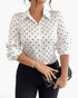 A woman wearing a white long-sleeve button-up blouse with black polka dots, paired with black pants and holding a black clutch bag with a gold chain strap, against a plain white background.