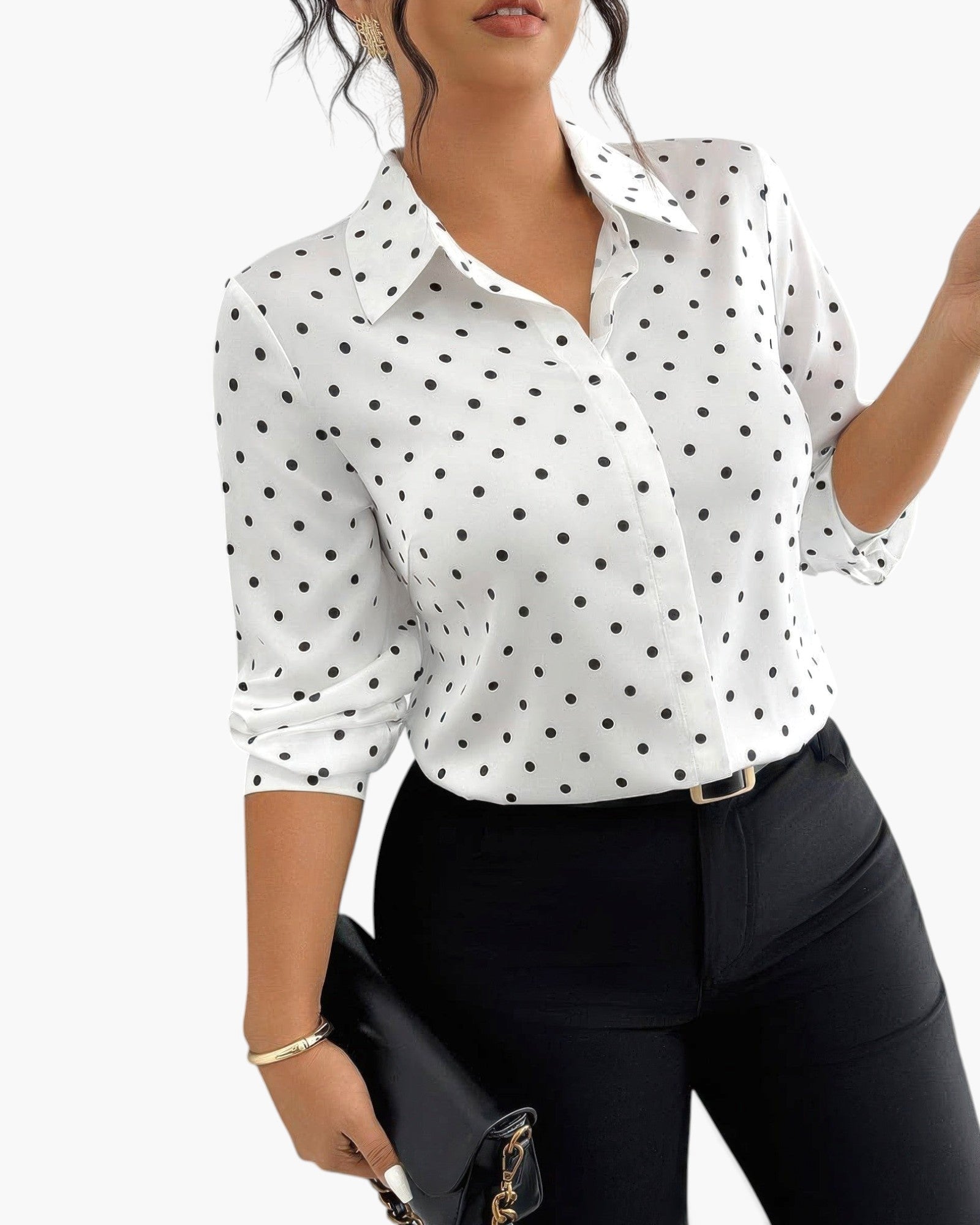 A woman wearing a white long-sleeve button-up blouse with black polka dots, paired with black pants and holding a black clutch bag against a plain white background.