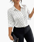 A woman wearing a white long-sleeve button-up blouse with black polka dots, paired with black pants and holding a black clutch bag against a plain white background.