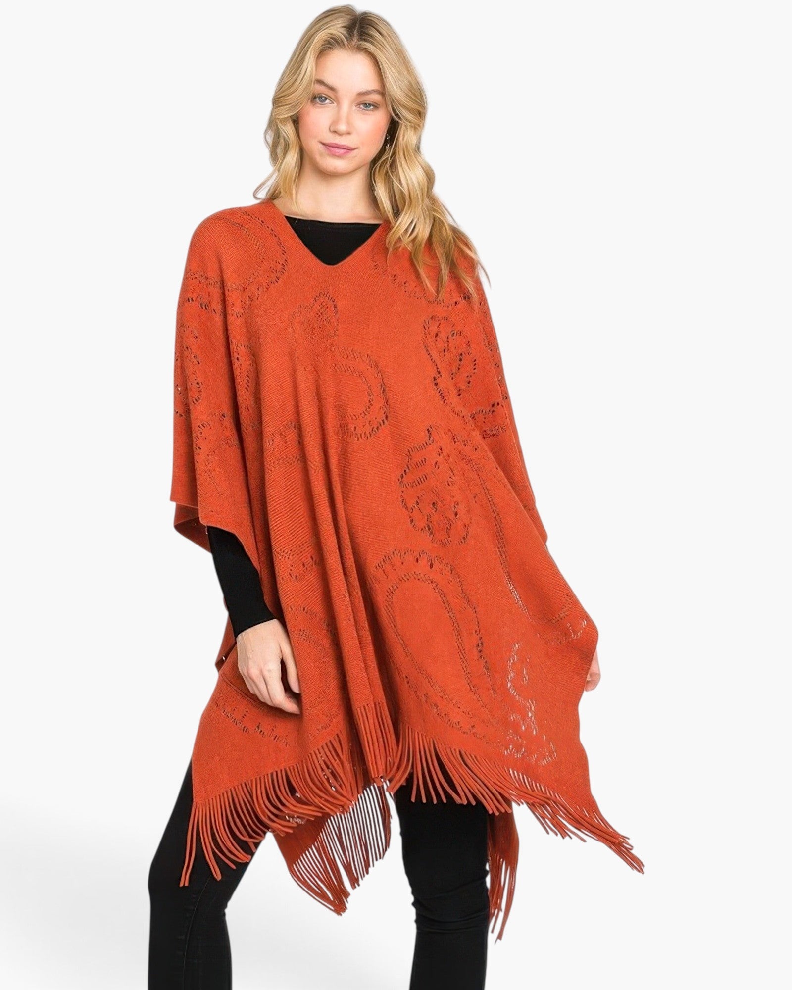 Beach Rose Co. model wearing a rust orange acrylic poncho with a paisley pattern and fringe hem over black clothing.