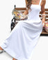 Beach Rose Co. white sleeveless A-line maxi dress with a square neckline, worn by a woman sitting on a park bench.