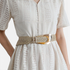 Beach Rose Co. beige woven polyester belt with an oversized gold alloy prong buckle worn over a white lace dress.