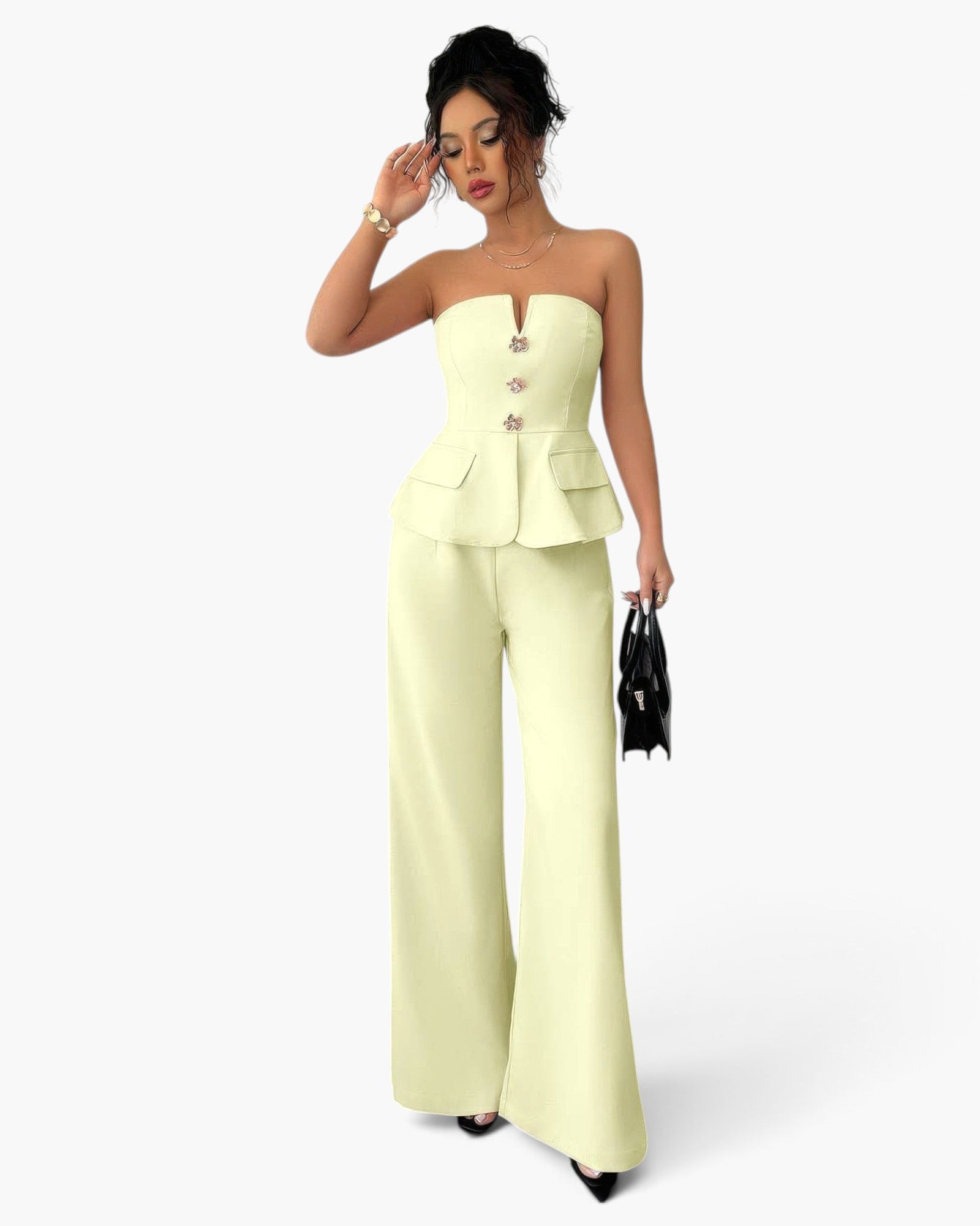 A woman models a pale yellow strapless set with a structured corset-style bodice, front buttons, and wide-leg pants, standing against a light gray studio background and holding a small black handbag.