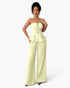 A woman modeling a pale yellow strapless set with a structured corset-style bodice, decorative front buttons, a peplum waist, and wide-leg pants, photographed against a light gray studio background.