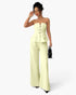 A woman models a pale yellow strapless set with a structured corset-style bodice, front buttons, and wide-leg pants, standing against a light gray studio background and holding a small black handbag.