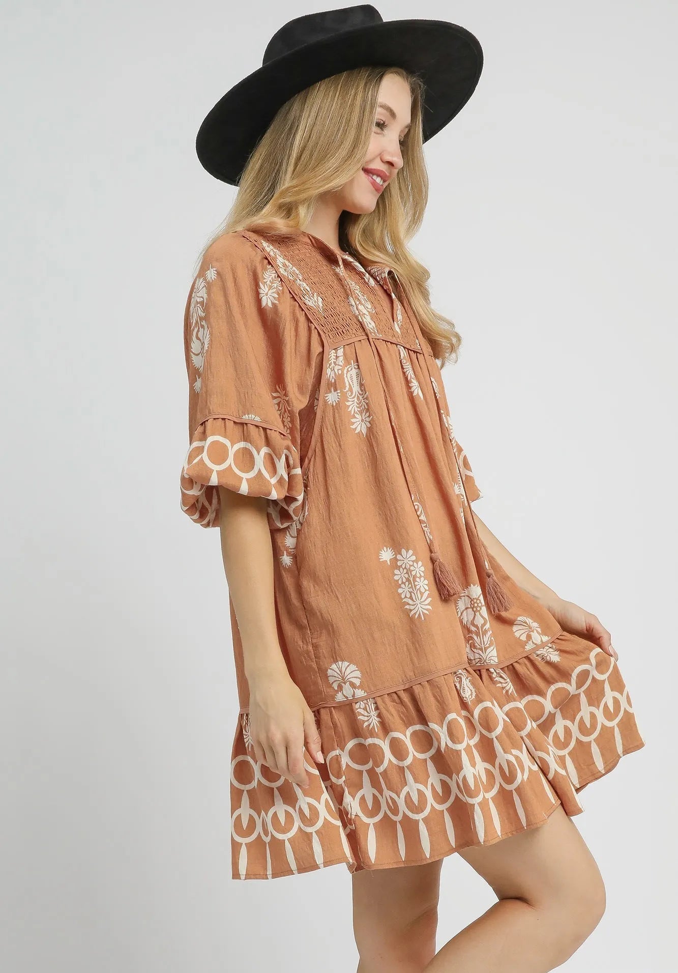 Woman wearing a brown dress with white patterns and a black hat on a light gray background