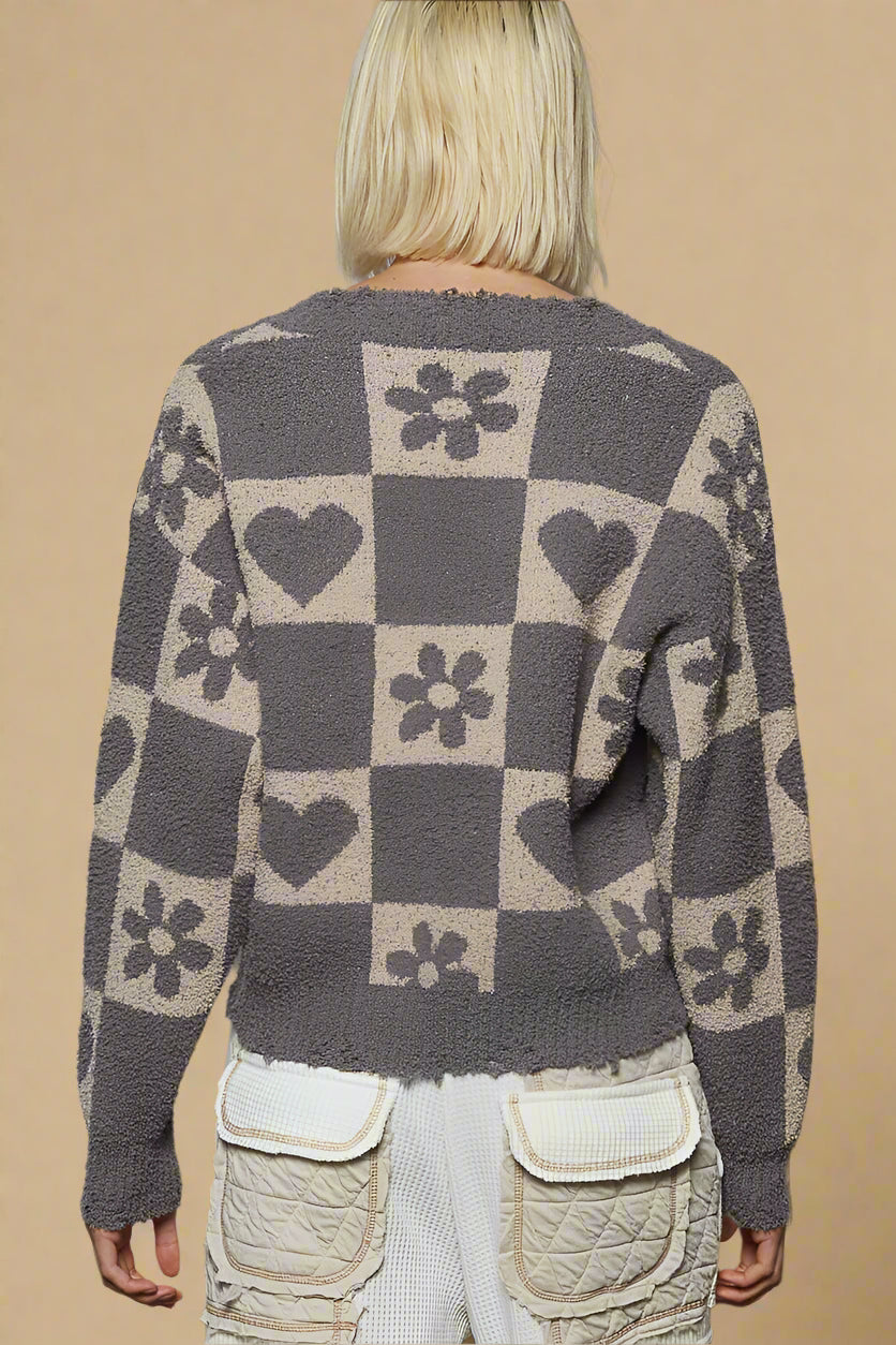 Person wearing a patterned sweater with hearts, flowers, and checkered design against a plain background