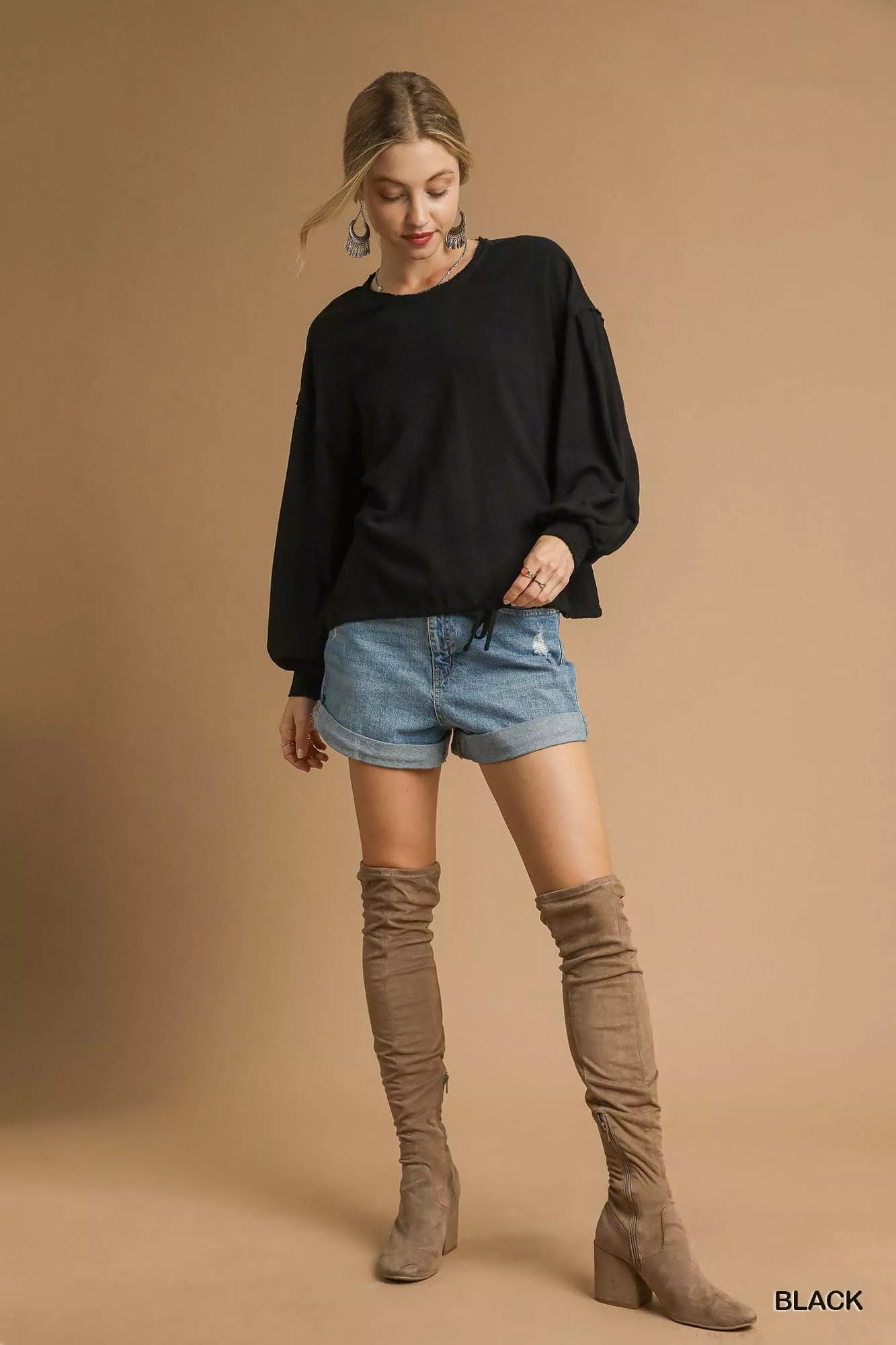 Woman wearing a black sweater, denim shorts, and brown knee-high boots on a beige background