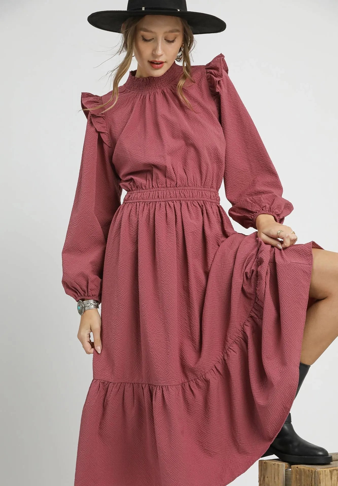 Woman wearing a dusty rose dress with ruffled sleeves and a black hat on a white background