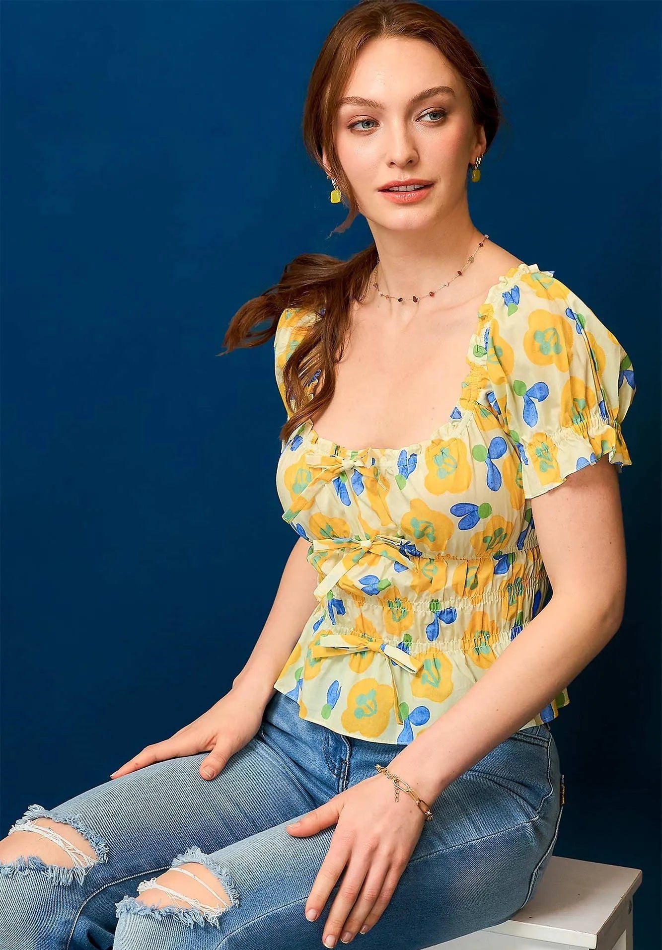 Woman wearing a yellow floral top and blue jeans sitting on a white stool against a blue background