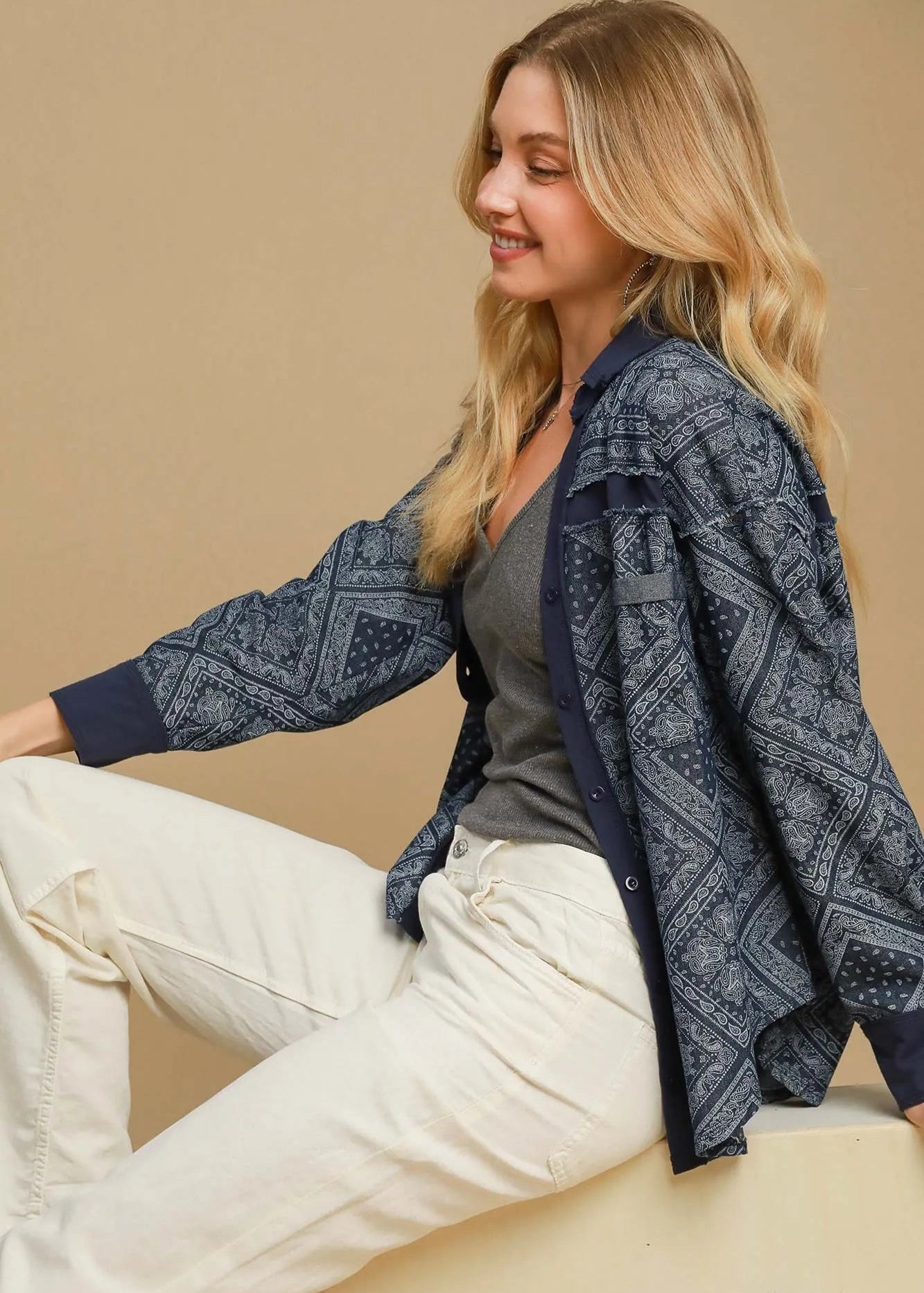 Woman wearing a blue patterned cardigan over a gray top and white pants on a beige background