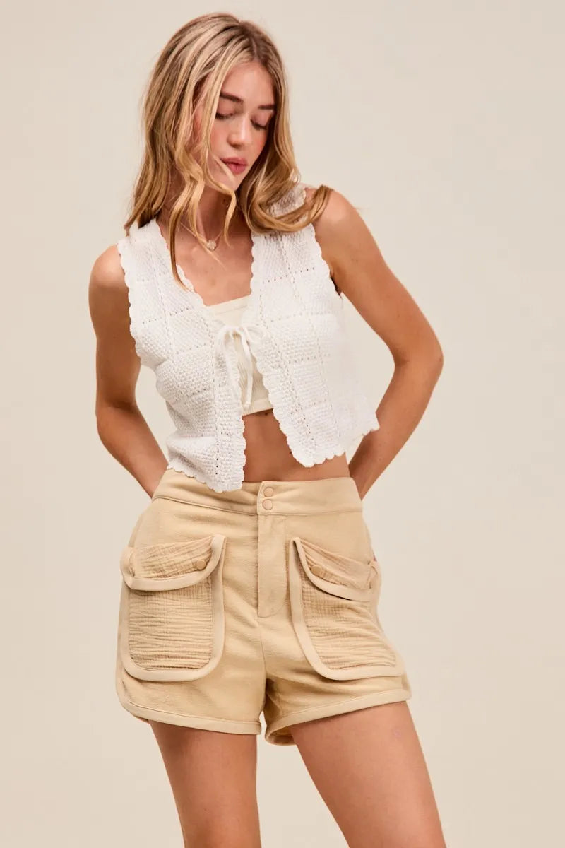 Woman wearing a white lace top and beige shorts on a plain background