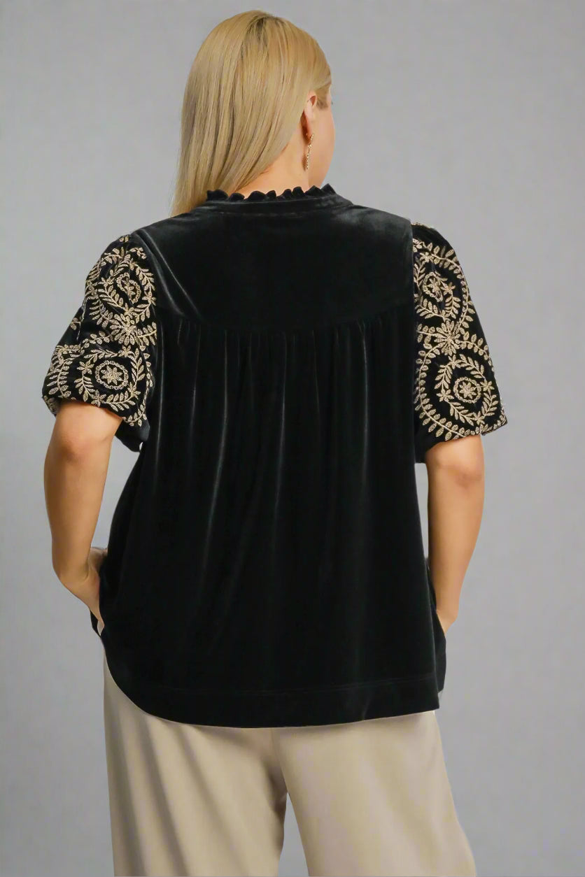 Black blouse with gold embroidery on sleeves worn by a model on a gray background
