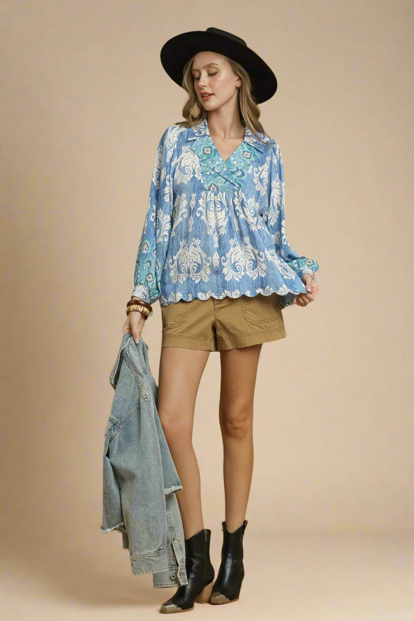 Woman wearing a blue patterned blouse, brown shorts, black boots, and a black hat on a beige background.