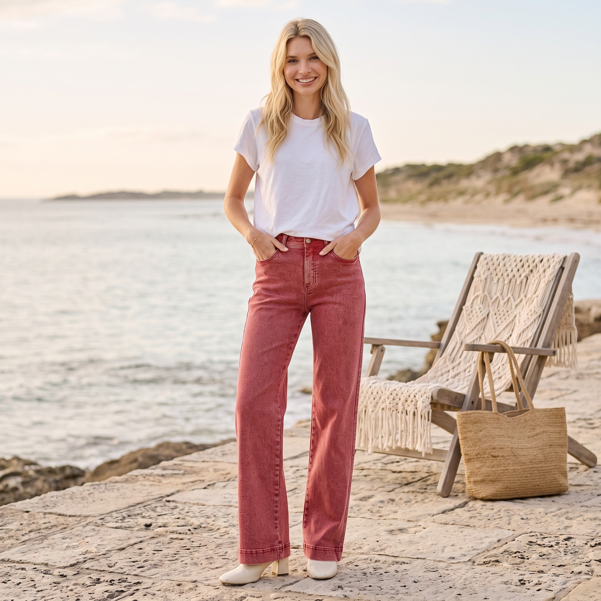 A woman stands on a stone beach walkway wearing a white t-shirt and dusty rose flare jeans, with a beach chair and ocean in the background.