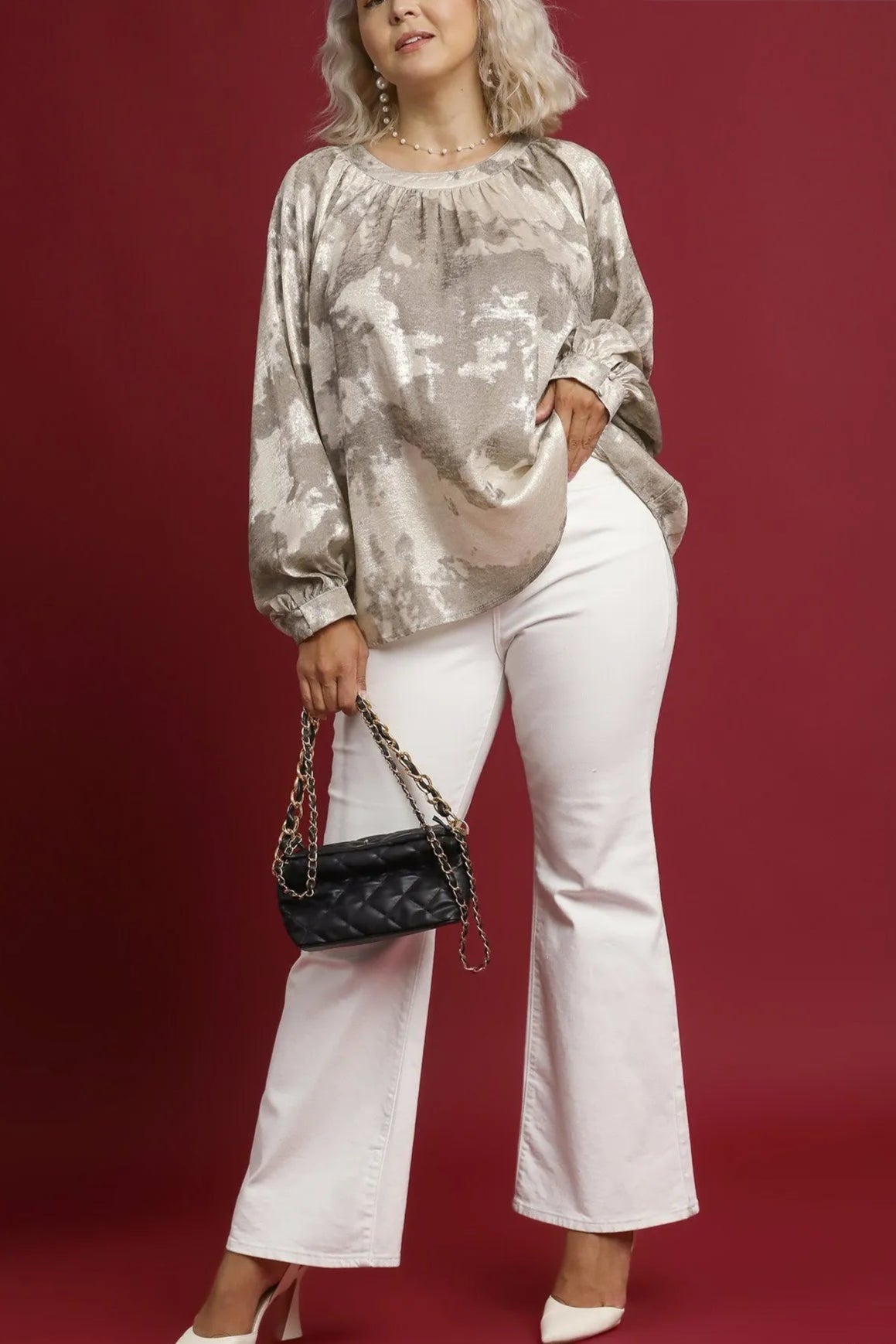 Woman wearing a taupe sequin top and white pants, holding a black handbag on a red background.