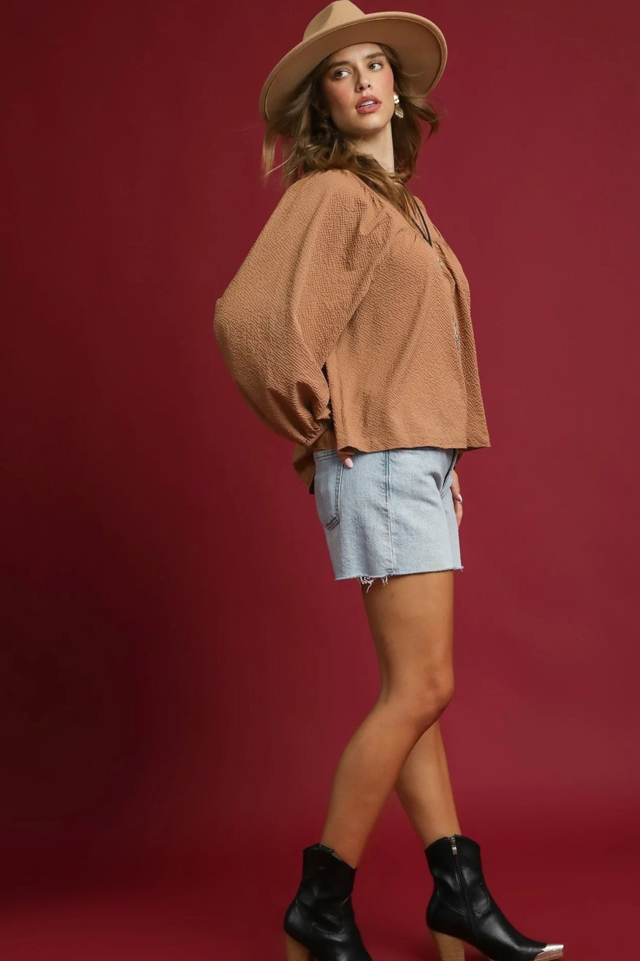 Woman wearing a brown sweater, light blue shorts, and black boots against a red background