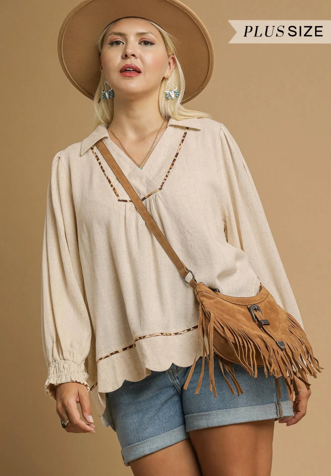 Woman wearing a beige blouse, denim shorts, and a brown hat with a fringe bag on a beige background.