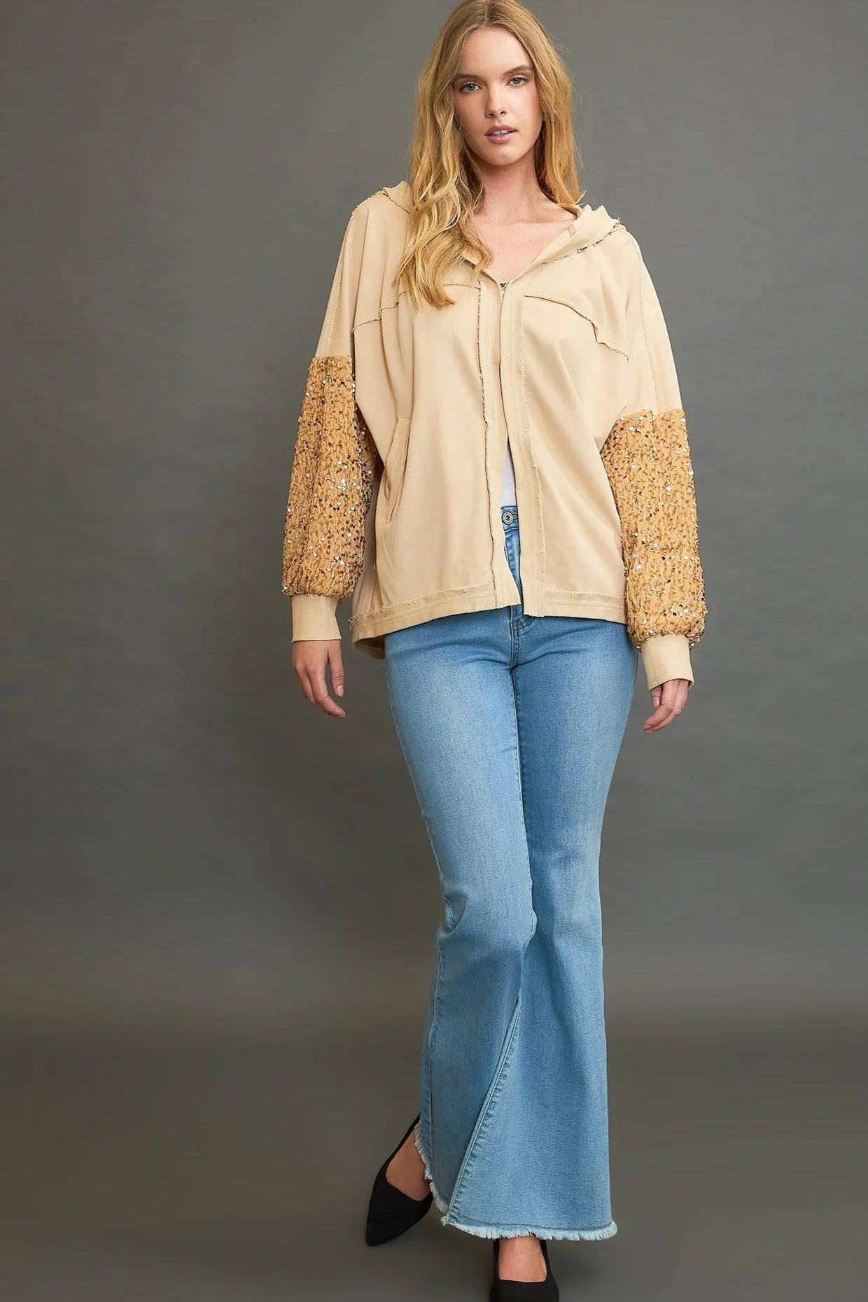 Woman wearing a beige jacket with textured sleeves and blue jeans on a gray background