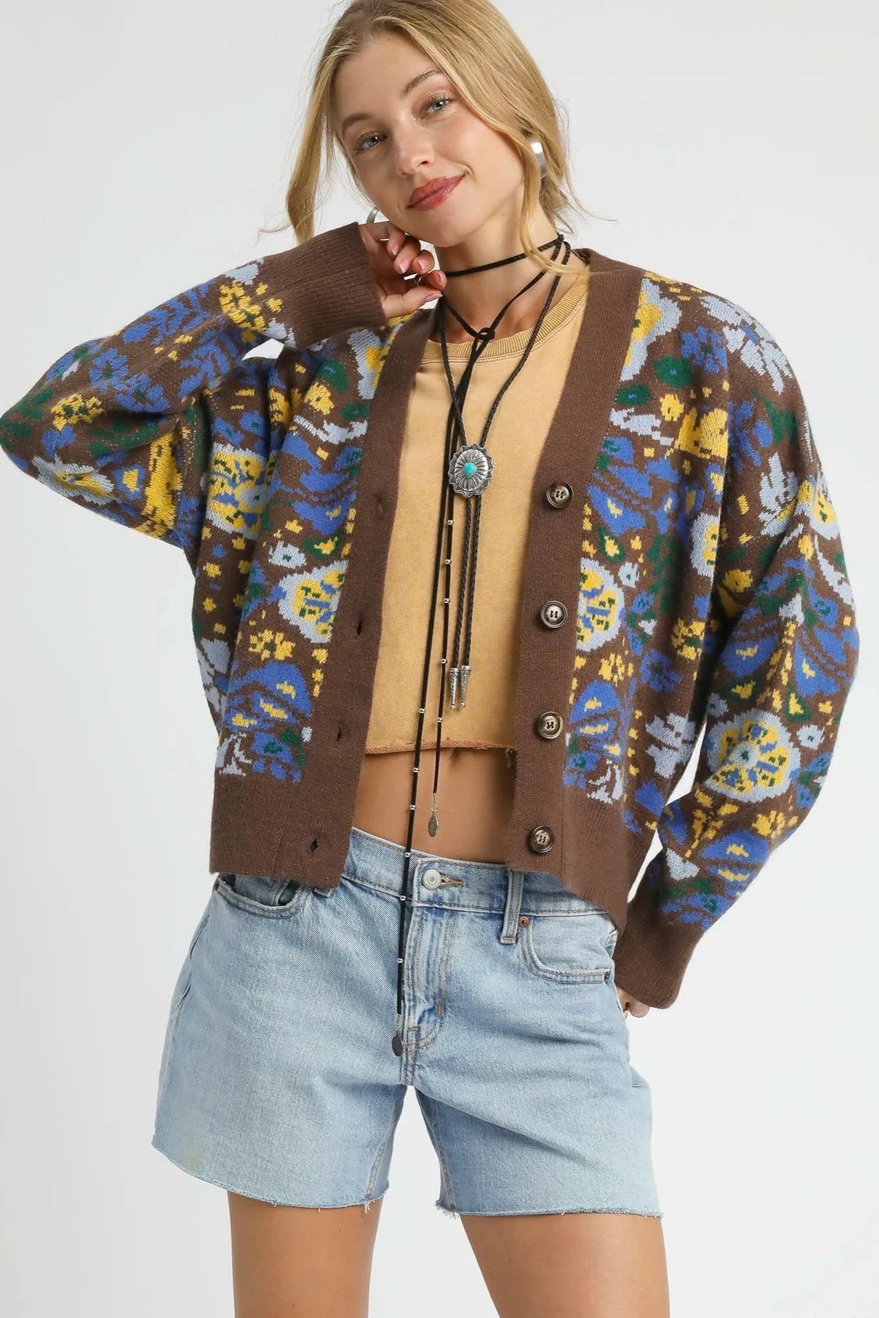 Woman wearing a patterned cardigan over a beige top and denim shorts on a white background